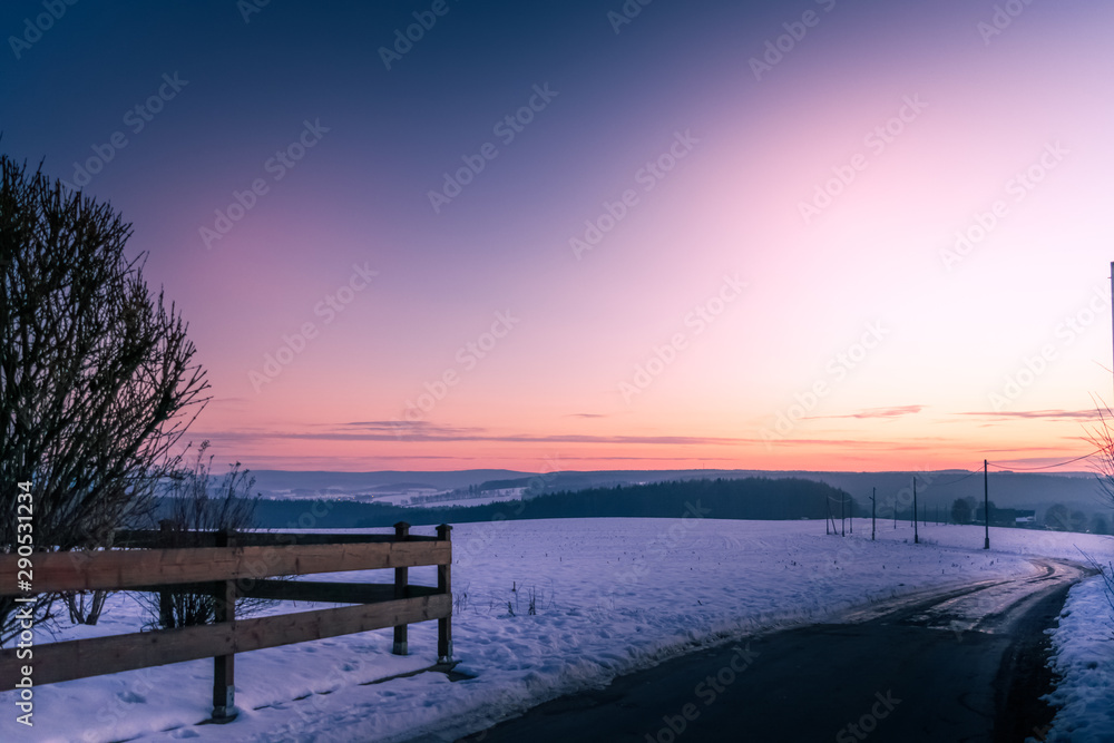 Colorful purple and orange sunset over a snowy landscape with rustic wooden fence and winding road over rolling hills
