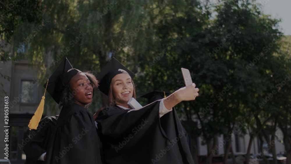 Selfie at graduation. Cheerful multi-ethnic girls posing to smartphone camera while funny classmates suddenly take a part in group photo. Mantle, academic hats, diploma. University on the background. Selfie at graduation. Cheerful multi-ethnic girls posing to smartphone camera while funny classmates suddenly take a part in group photo. Mantle, academic hats, diploma. University on the background.