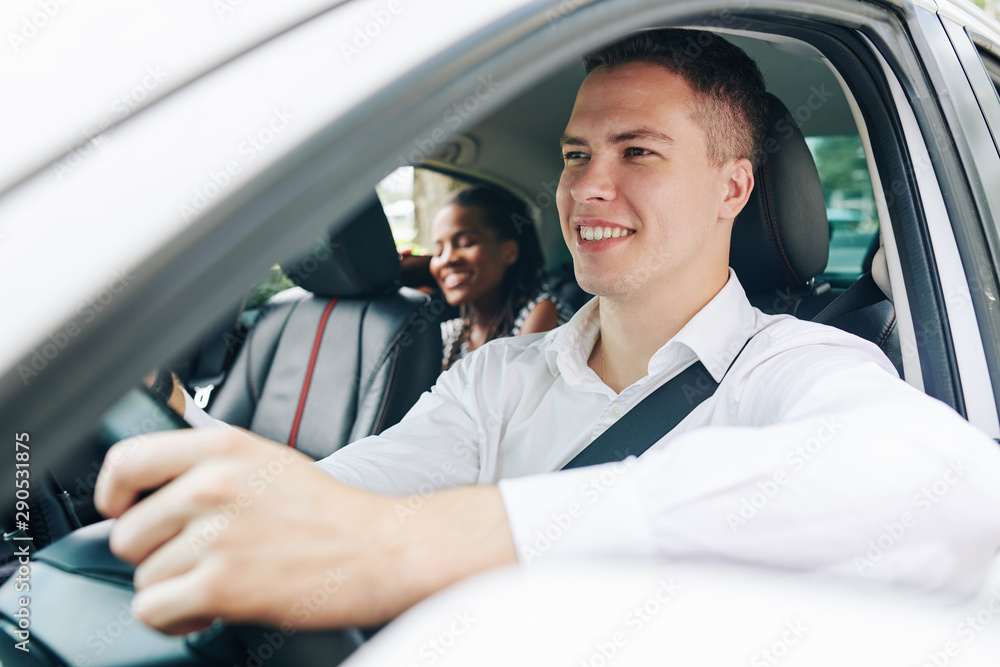 Young smiling man holding the wheel and driving a car with young woman sitting on back seat