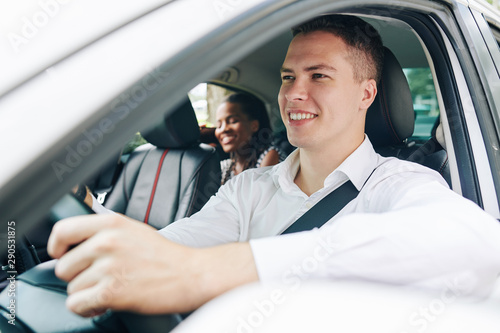 Young smiling man holding the wheel and driving a car with young woman sitting on back seat