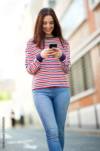 smiling young woman walking in city and looking at cellphone © mimagephotos