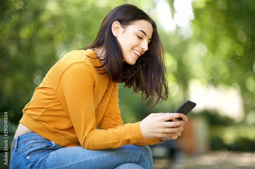 young woman smiling and looking at mobile phone while sitting on park bench