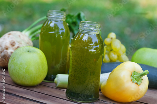 Healthy green smoothie with apples, grapes, cucumber and celery. Fruit and vegetable juice in little bottles on wooden table, with blured nature in background.