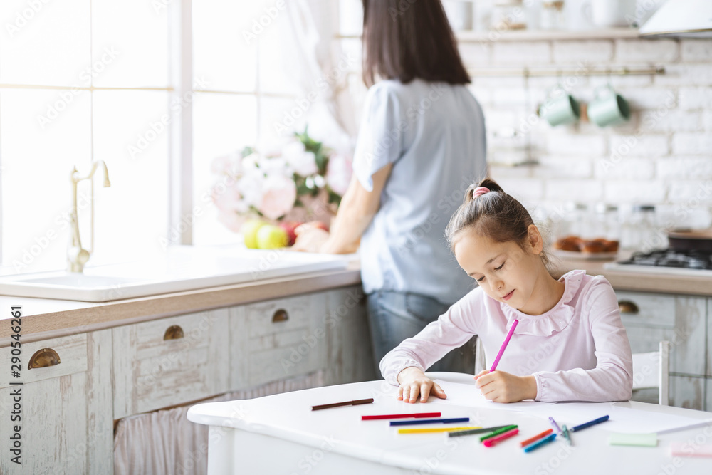 Cute girl drawing at kitchen table while her mother cooking Cute girl drawing at kitchen table while her mother cooking