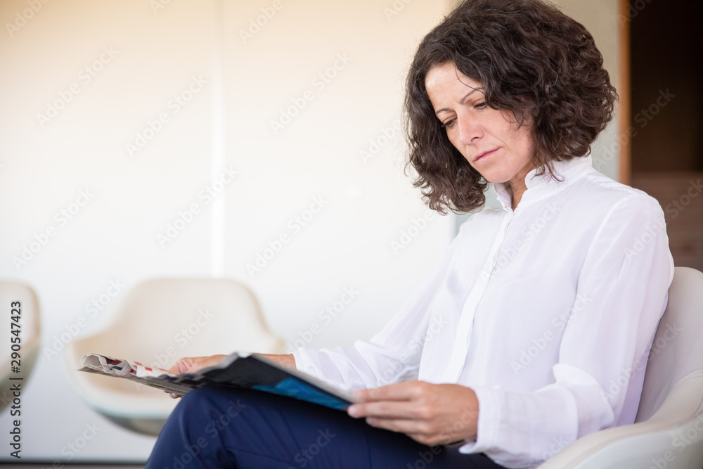 Serious female office worker reading magazine during work break. Business woman sitting in armchair and holding newspaper in office lounge. News concept Serious female office worker reading magazine during work break. Business woman sitting in armchair and holding newspaper in office lounge. News concept