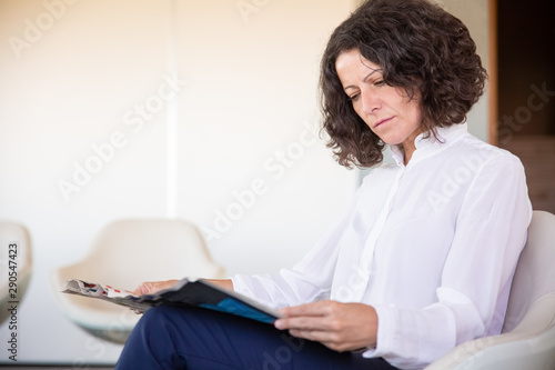 Serious female office worker reading magazine during work break. Business woman sitting in armchair and holding newspaper in office lounge. News concept