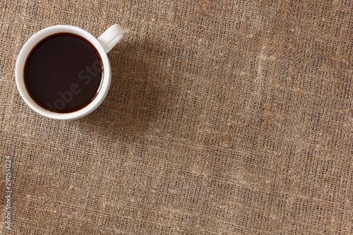 White mug with coffee on rough burlap tablecloth, top view.