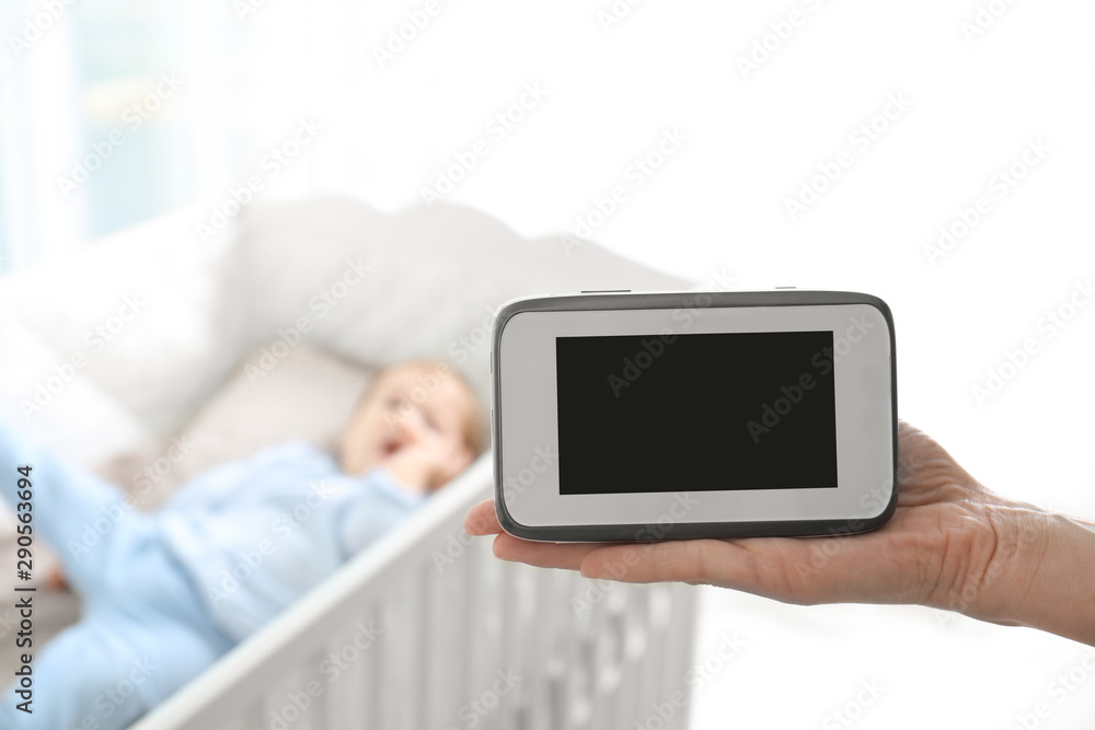 Woman holding baby monitor near crib with child in room. Video nanny
