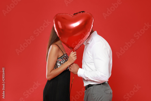 Couple kissing while hiding behind heart shaped balloon on red background