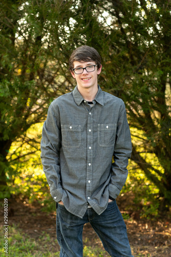 Teenage boy with glasses standing outside in front of a tree wearing a gray button up long sleeved shirt and denim jeans and smiling