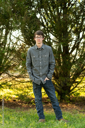 serious Teenage boy with glasses standing outside in front of a tree wearing a gray button up long sleeved shirt and denim jeans