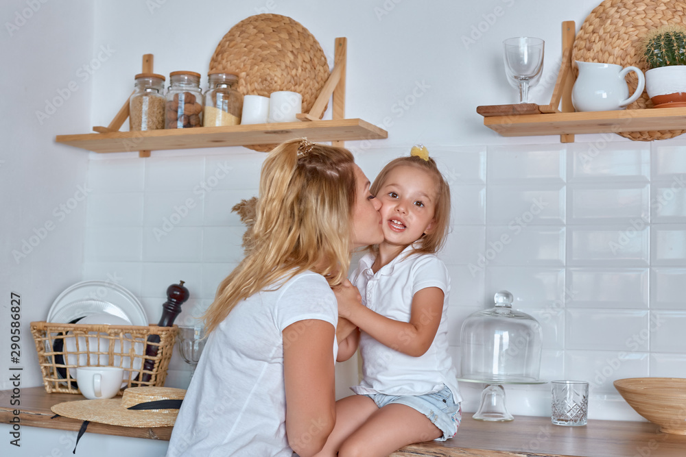Obraz premium Little girl with mom smile and laugh. A girl sits on the working surface of the kitchen. A cheerful and mischievous daughter with her mother spends time in the kitchen.