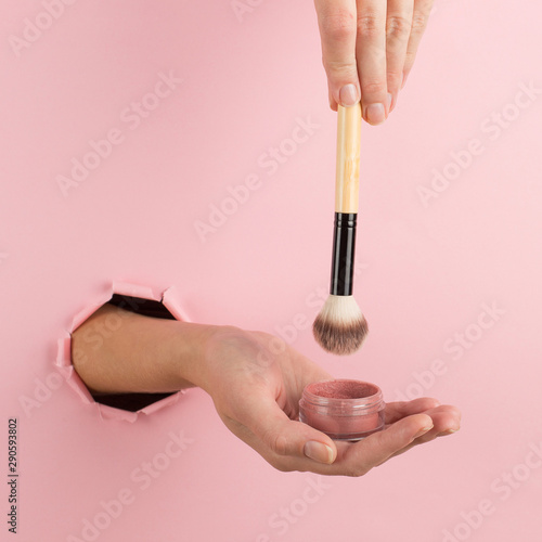 Girl hand holds a brush for makeup and powder from a hole in a pink background. Makeup artist concept, copy space.