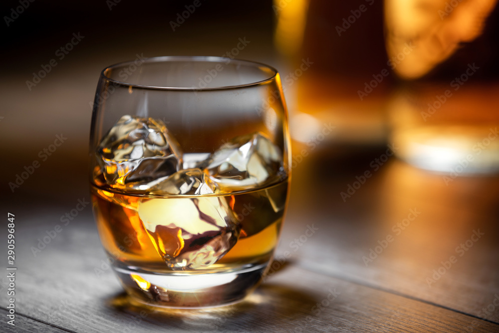 Elegant and refreshing glass of scotch bourbon whisky on ice with glowing illuminated golden bottles in background, copy space Elegant and refreshing glass of scotch bourbon whisky on ice with glowing illuminated golden bottles in background, copy space