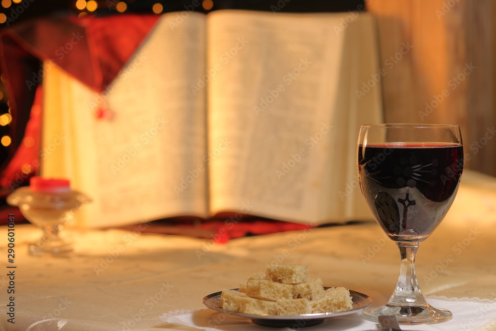 Concept Lord's Supper and Christian communion. Bread and wine in chalice are front of big Bible. Cross reflect in goblet, candle is close and light on background. Concept Lord's Supper and Christian communion. Bread and wine in chalice are front of big Bible. Cross reflect in goblet, candle is close and light on background.
