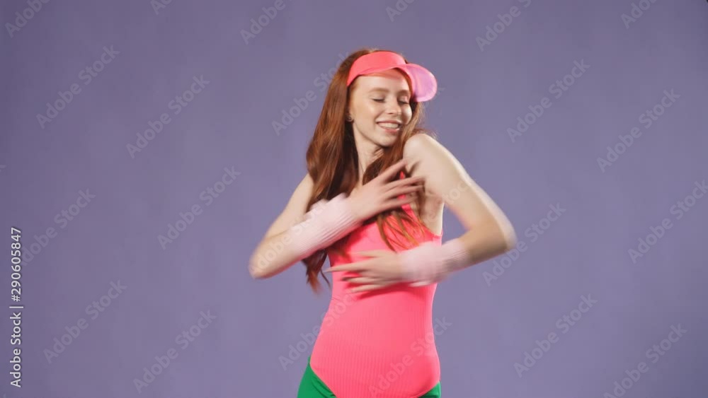Pretty young red-headed female aerobics instructor in pink bodysuit, green leggins and pink sun cap in retro style of 80s, stands relaxed and looks in camera Pretty young red-headed female aerobics instructor in pink bodysuit, green leggins and pink sun cap in retro style of 80s, stands relaxed and looks in camera