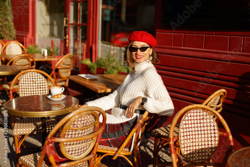 Photography Young beautiful elegant woman in French style cafe wearing in white sweater and red beret and eyeglasses, sitting in chair and with cup of coffee