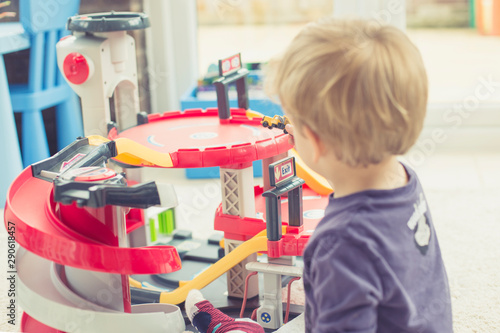 Blond toddler in purple t-shirt playing with his cars in a red toy parking in his room