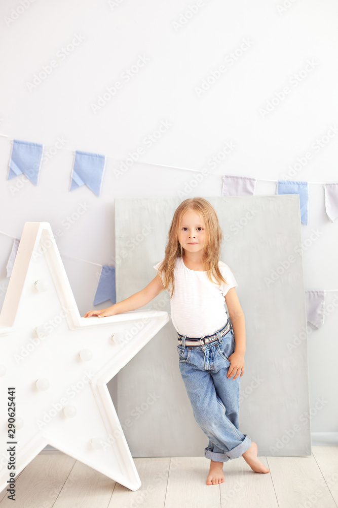 little girl in jeans and a white T-shirt stands by the luminous star of the lamp. Large decorative retro star. Christmas, New Year. Big star with light bulbs on a concrete wall. Scandinavian interior