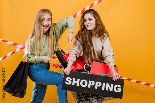 two happy smiling beautiful women have cart and shopping sign with colorful shopping bags and signal tape isolated over yellow photo