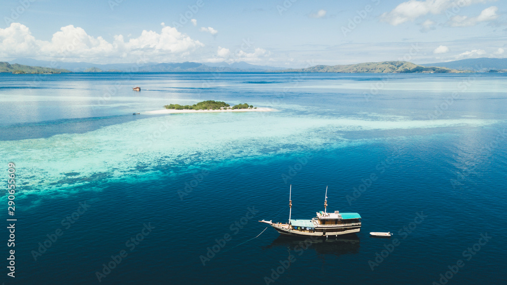 Fototapeta premium Luxury cruise boat sailing near coral reef atoll island with amazing white tropical beach and mountains on horizon. Aerial view. Luxury marine travel and vacations concept.