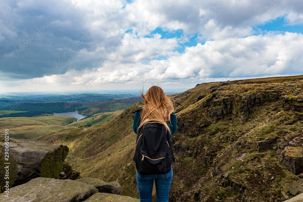 A woman with blonde hair wearing a blue woolen jumper, blue jeans and a black backpack on her, stands on a rock to take a photo of the rolling hills in the Peak District on a cloudy day in England, UK