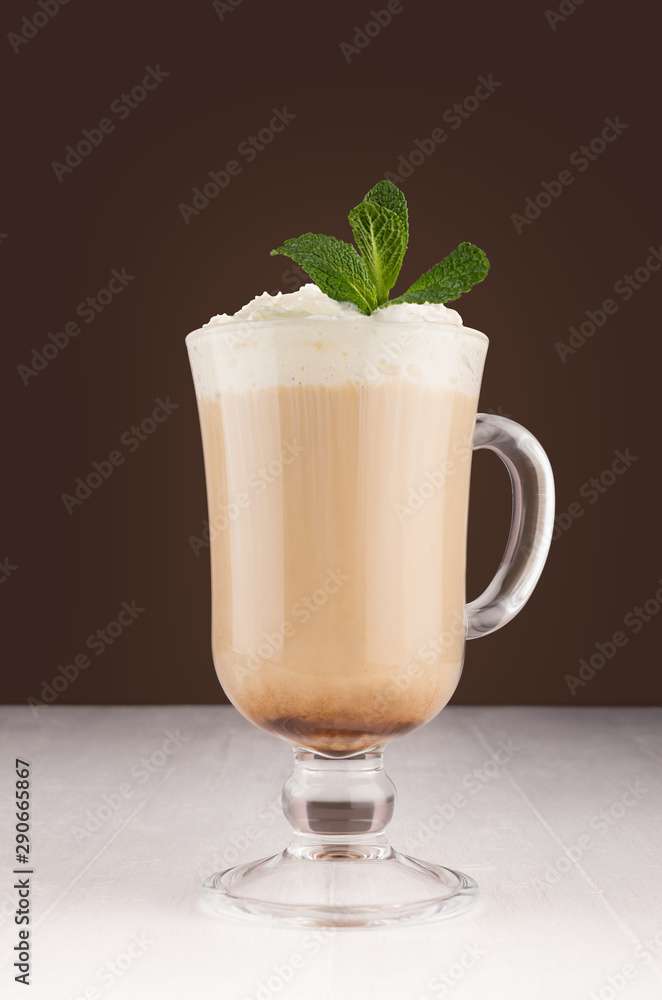 Coffee drink with cream and fresh green mint for breakfast in transparent glass with handle in dark brown interior on white wood table. Coffee drink with cream and fresh green mint for breakfast in transparent glass with handle in dark brown interior on white wood table.