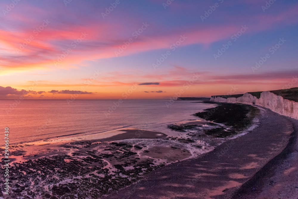 Obraz premium A sun sets over the clouds and paints golden and pink colours across the skies as the rocky shore and the white chalk cliffs of the Seven Sisters meets the sea in Eastbourne, UK.