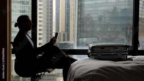Man sitting in chair at dark room against window, looking to cell phone, super slow motion shot. Traveller arrive to destination, take pause after long flight. Packed suitcase lie on bed
