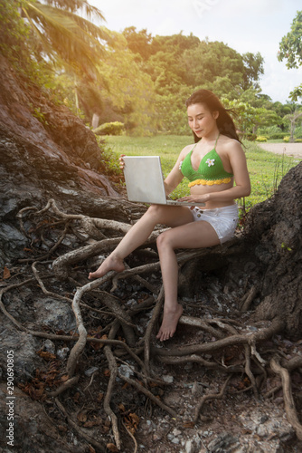Canvas Print A beautiful Asian woman sitting and working with a laptop  at the root of a tree on  large rock