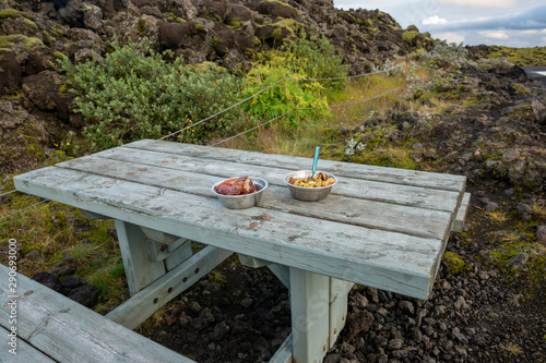 Metal bowls with lamb steaks and potatoes on wooden picnic table. Stone landscape, Iceland.