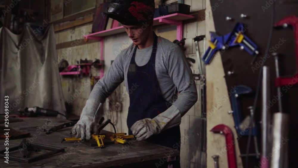 Tracking medium shot of young metalworker in protective gloves and mask gripping metal construction with vises and welding it with welding torch in slow motion standing at workbench Tracking medium shot of young metalworker in protective gloves and mask gripping metal construction with vises and welding it with welding torch in slow motion standing at workbench
