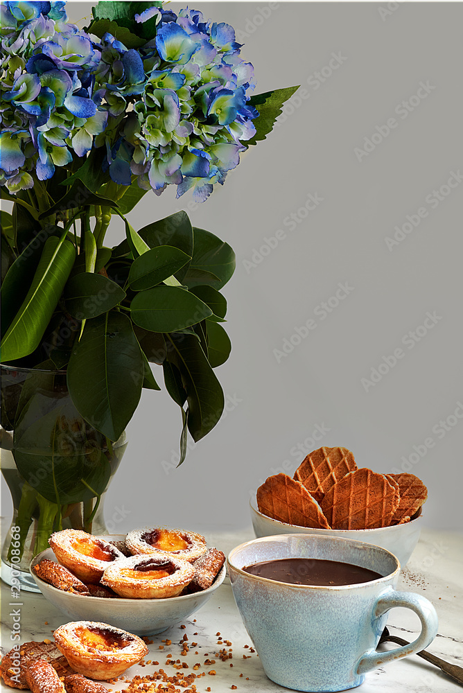 Cup of homemade chocolate accompanied by puff pastry cakes filled with custard and cookies. In the background a blue flowers.