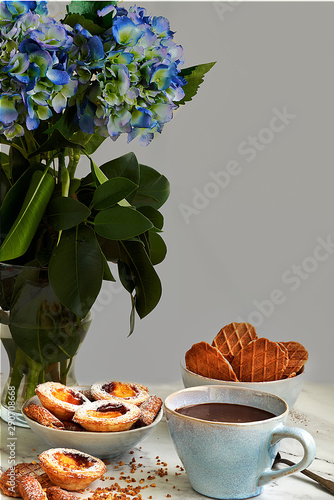 Obraz na płótnie Cup of homemade chocolate accompanied by puff pastry cakes filled with custard and cookies. In the background a blue flowers. w salonie