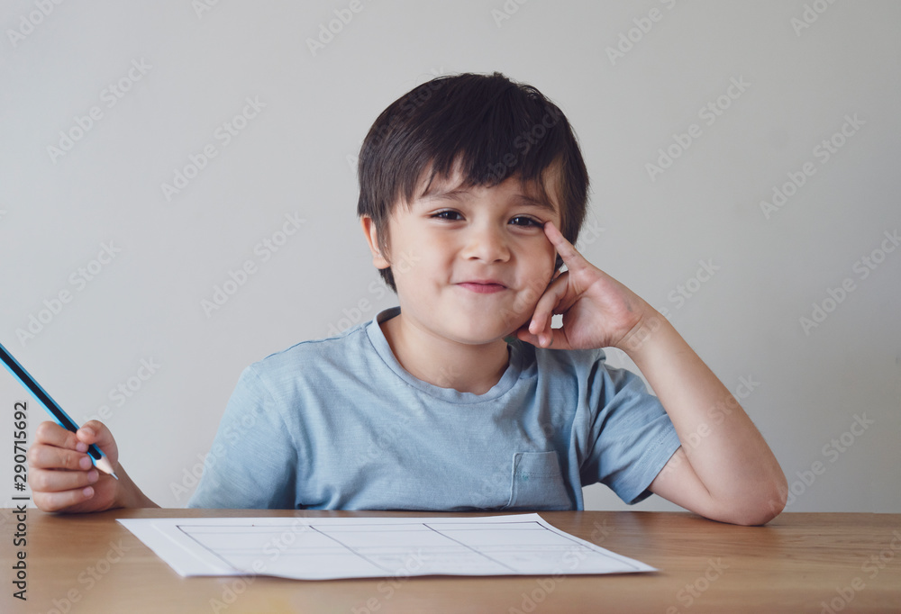 Portrait of preschool kid boy making homework at home. Happy Child holding pencils and looking at camera with smiling face, Little boy is writing at the table, Elementary school and education concept Portrait of preschool kid boy making homework at home. Happy Child holding pencils and looking at camera with smiling face, Little boy is writing at the table, Elementary school and education concept