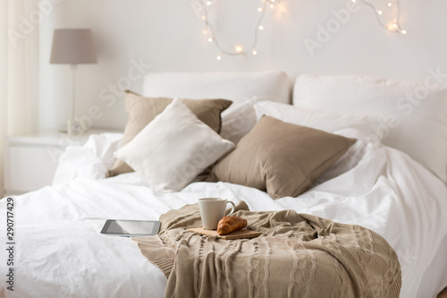 comfort, hygge and interior concept - tablet computer, cup of coffee and croissant on bed at cozy home