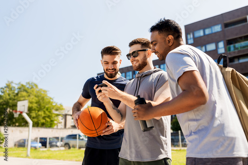 Fototapeta Naklejka Na Ścianę i Meble -  sport, leisure games and male friendship concept - group of men or friends with smartphone at outdoor basketball playground