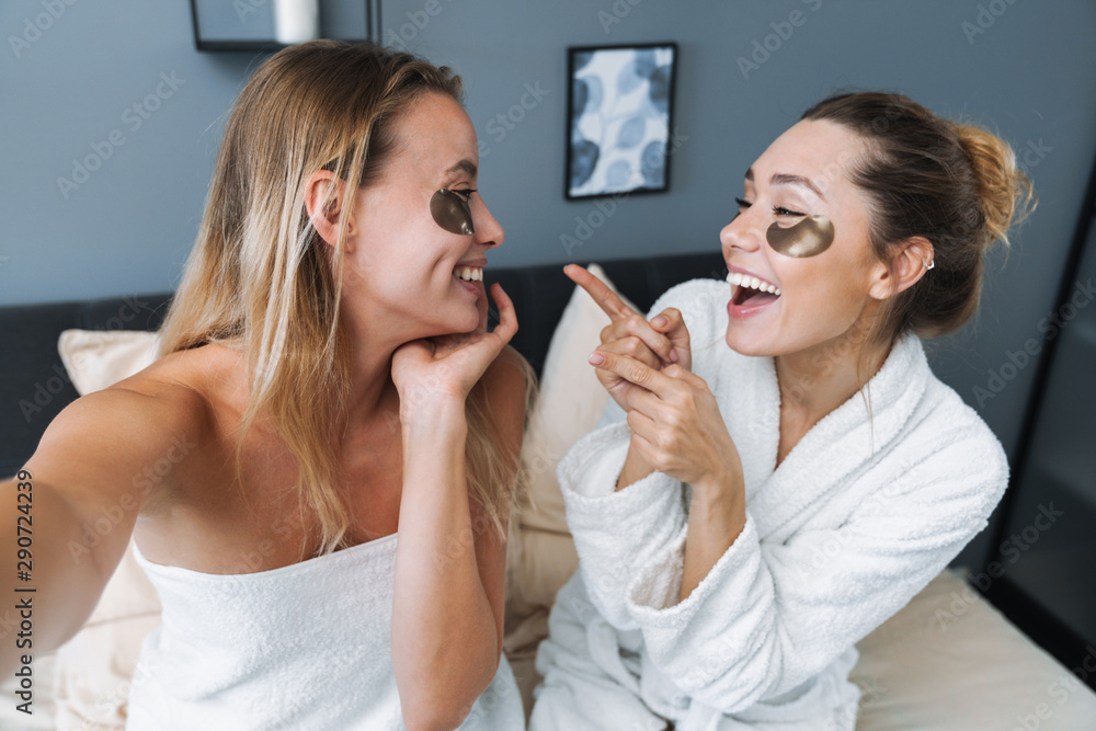 Pleased young women taking care of their skin with under eye patches. Pleased young women taking care of their skin with under eye patches.