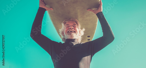 happy senior with surftable on his head is smiling and laughing - old and mature man having fun surfing with a black wetsuits - active retired adult doing activity alone photo