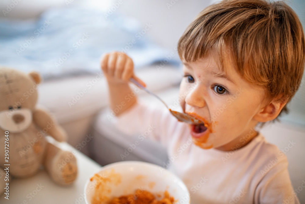 Eating cheerful baby boy with messy face. Portrait of a toddler eating. Open mouth child with an appetite eats millet porridge. Happy cute baby kid eating food itself with spoon. Eating cheerful baby boy with messy face. Portrait of a toddler eating. Open mouth child with an appetite eats millet porridge. Happy cute baby kid eating food itself with spoon.