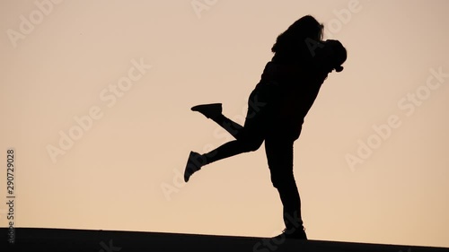 Man hug woman tight and rise up from ground, happy girl wave legs in air. Silhouette of lovely pair against dim yellow sky. Couple embrace one another and kiss, stay close together photo
