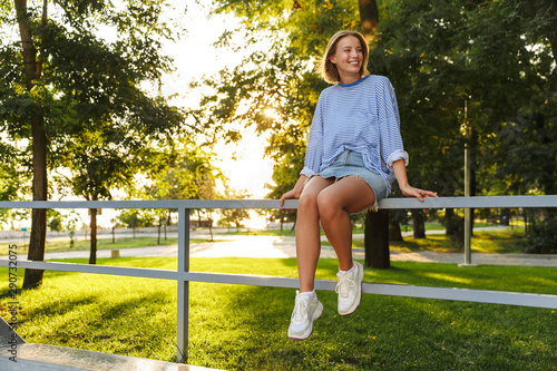 Image of romantic happy woman smiling and sitting on railing in park