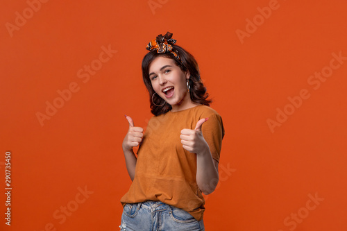 Beautiful brunette woman in an orange t-shirt and headband holding thumbs up expressing positive evaluation standing isolated over orange background. I like it!