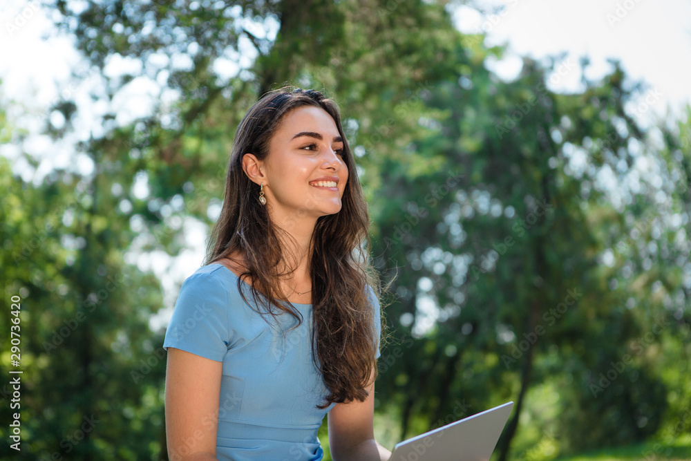 Young brunette student in a blue dress sitting with a laptop in a park. Distance learning Young brunette student in a blue dress sitting with a laptop in a park. Distance learning