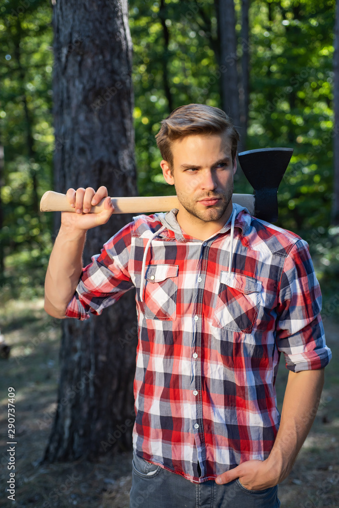 Obraz premium Stylish young man posing like lumberjack. The Lumberjack working in a forest. Handsome man with axe. Strong man lumberjack with an ax in a plaid shirt.