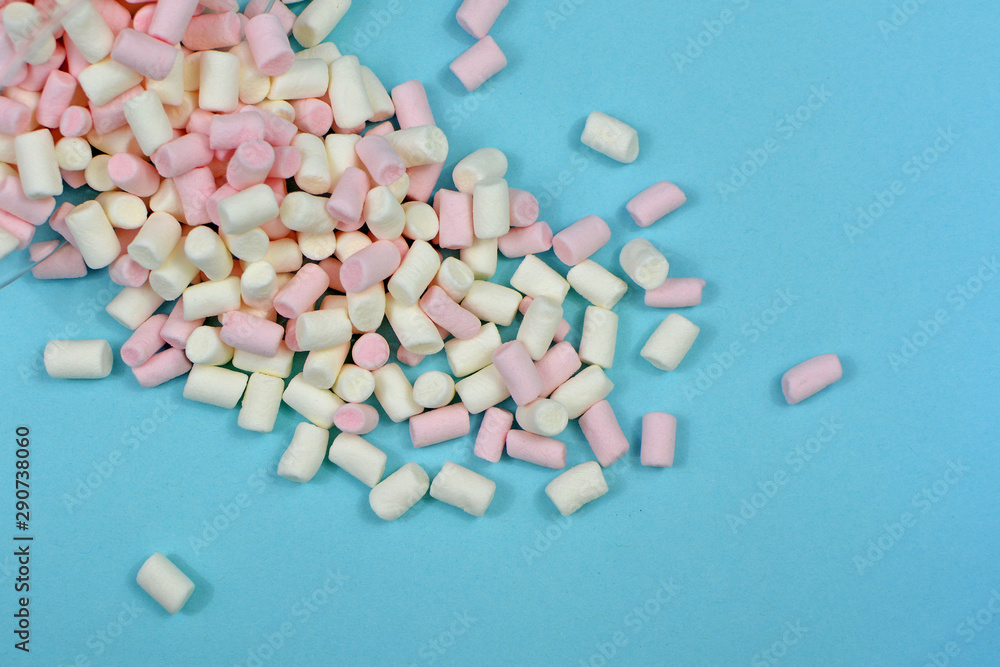 Pink and white small marshmallow weets spilling out of glass container on blue background Pink and white small marshmallow weets spilling out of glass container on blue background