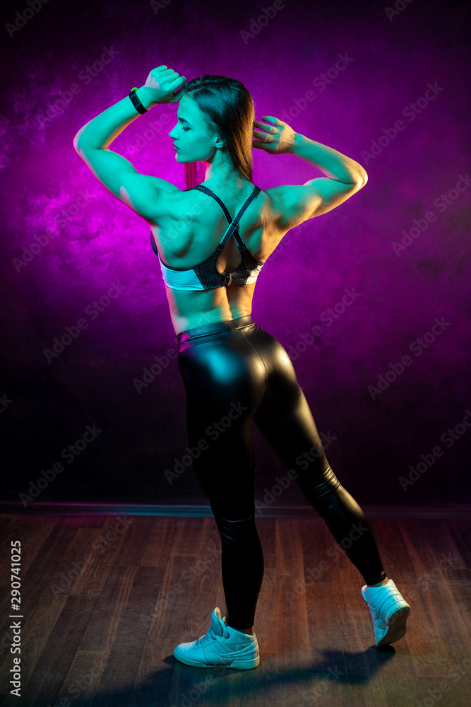 Full lenght photo of beautiful young woman fitness model posing in neon lights in the studio. Sporty woman show her back muscles.