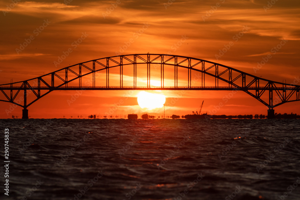 Fototapeta premium Steel tied arch bridge over water with a sunset behind it. Telephoto large sun - Fire Island Inlet Bridge, Long Island. 