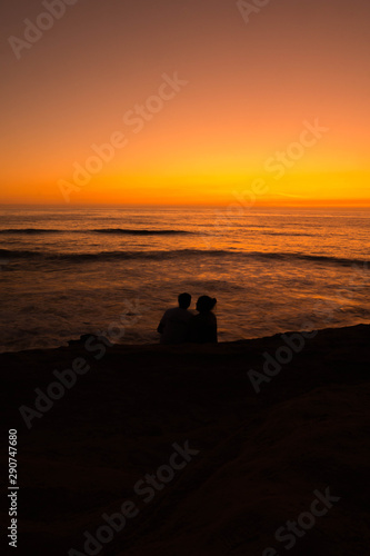 silhouette of a couple sitting on a cliff during sunset by the ocean