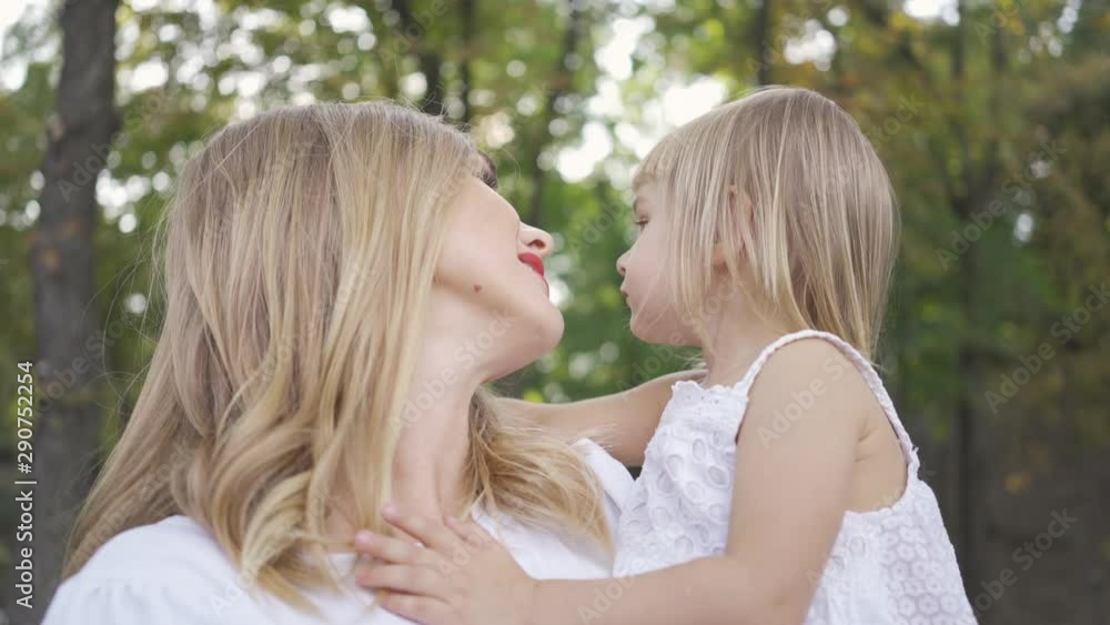 Young blond woman in white caressing her cute daughter. Daughter hugging her mother. Leisure outdoors. Happy family. One parent. Motherhood, parenthood, happy childhood Young blond woman in white caressing her cute daughter. Daughter hugging her mother. Leisure outdoors. Happy family. One parent. Motherhood, parenthood, happy childhood